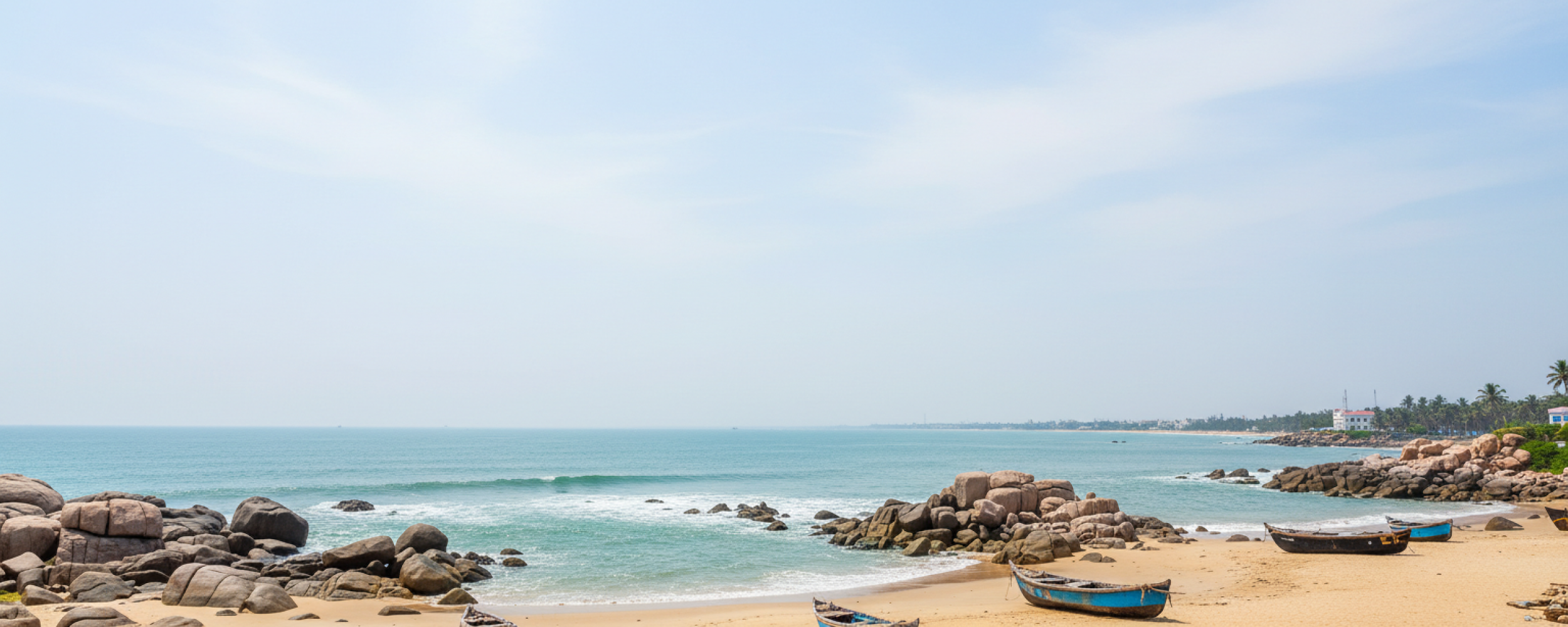 A tranquil sandy beach with wooden fishing boats, rocky outcrops, and calm turquoise waters — a perfect destination for day trips from Pondicherry