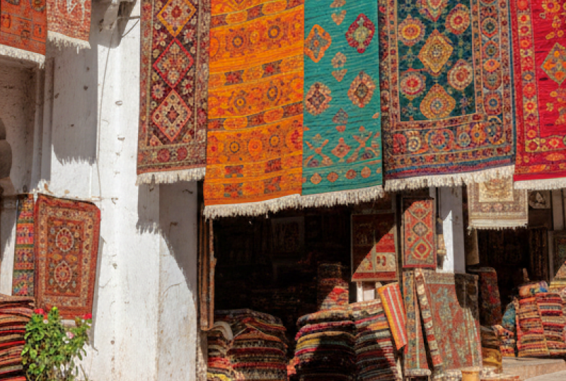 Colourful hand woven rugs and carpets hanging over a sunlit market lane, with stacked textiles outside the stalls, a popular find when shopping in Goa thumbnail