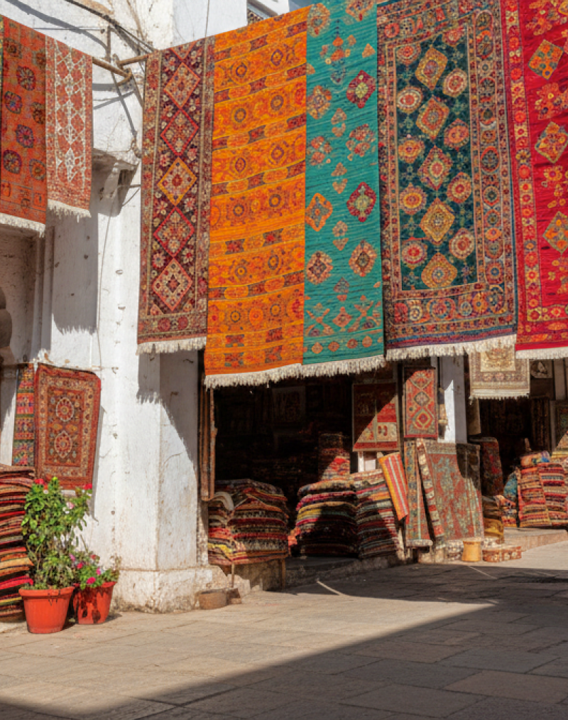 Colourful hand woven rugs and carpets hanging over a sunlit market lane, with stacked textiles outside the stalls, a popular find when shopping in Goa thumbnail