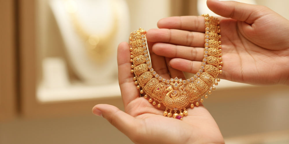 Hands holding an intricate traditional gold necklace with peacock motif and ruby accents, representing the experience of buying gold in India