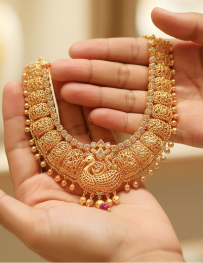 Hands holding an intricate traditional gold necklace with peacock motif and ruby accents, representing the experience of buying gold in India thumbnail