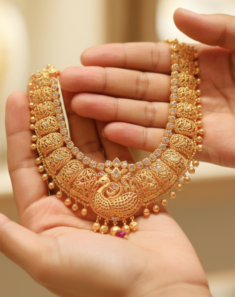 Hands holding an intricate traditional gold necklace with peacock motif and ruby accents, representing the experience of buying gold in India thumbnail