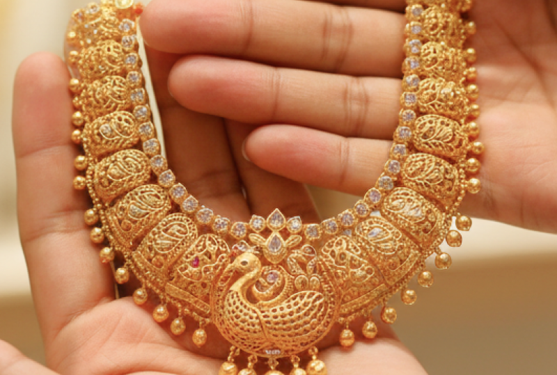 Hands holding an intricate traditional gold necklace with peacock motif and ruby accents, representing the experience of buying gold in India thumbnail