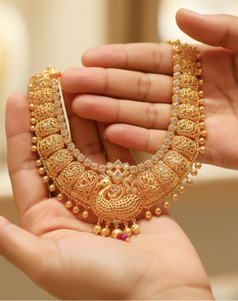 Hands holding an intricate traditional gold necklace with peacock motif and ruby accents, representing the experience of buying gold in India thumbnail