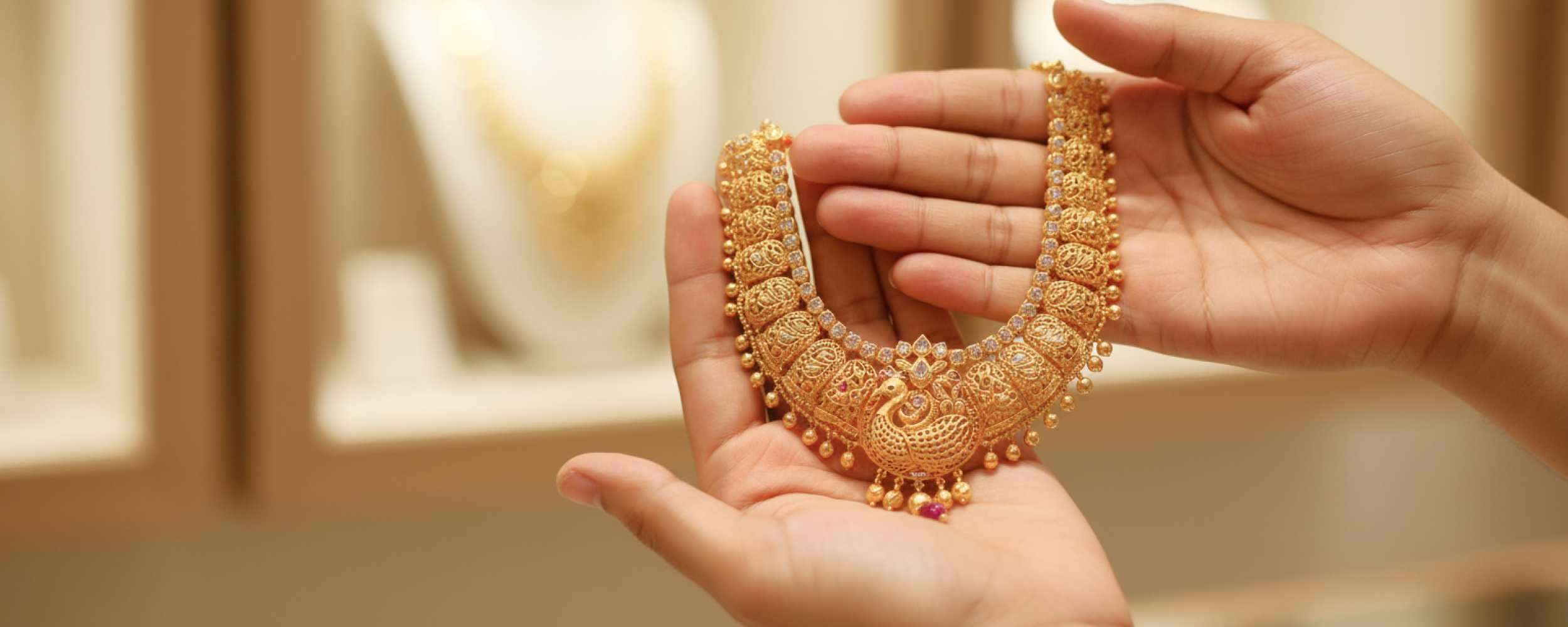 Hands holding an intricate traditional gold necklace with peacock motif and ruby accents, representing the experience of buying gold in India