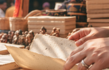 A shopper browsing handmade paper notebooks at a vibrant outdoor market stall in Pondicherry, India, with colourful sarees and craft goods visible in the background thumbnail