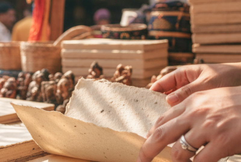 A shopper browsing handmade paper notebooks at a vibrant outdoor market stall in Pondicherry, India, with colourful sarees and craft goods visible in the background thumbnail