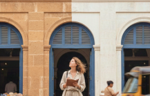 Female traveler standing outside historic colonial building with blue shutters, street market stalls, scooter and auto rickshaw in India thumbnail