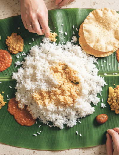 Overhead view of a traditional South Indian banana leaf meal (saapadu) with rice, chutneys, papad and fried fish, with two pairs of hands eating, served on a stone floor in Pondicherry thumbnail