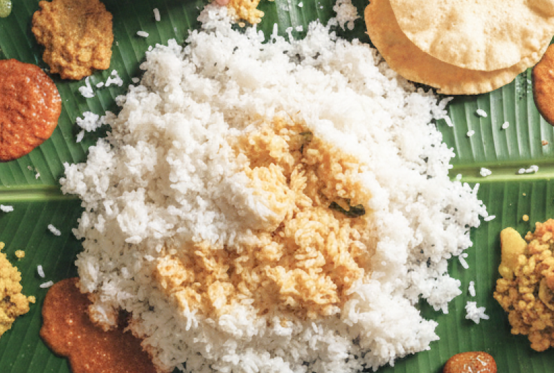 Overhead view of a traditional South Indian banana leaf meal (saapadu) with rice, chutneys, papad and fried fish, with two pairs of hands eating, served on a stone floor in Pondicherry thumbnail