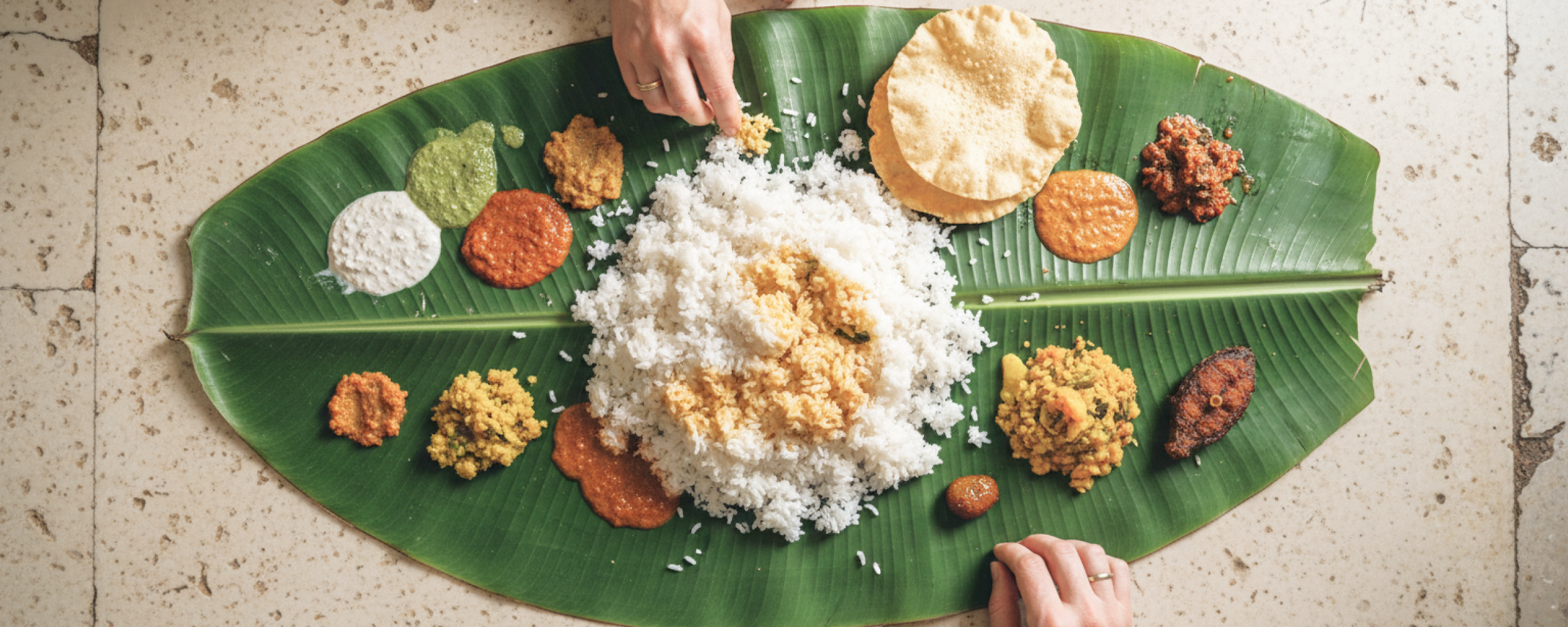Overhead view of a traditional South Indian banana leaf meal (saapadu) with rice, chutneys, papad and fried fish, with two pairs of hands eating, served on a stone floor in Pondicherry