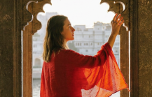 Woman in red shawl standing in ornate Indian palace window overlooking Lake Pichola in Udaipur at sunset thumbnail