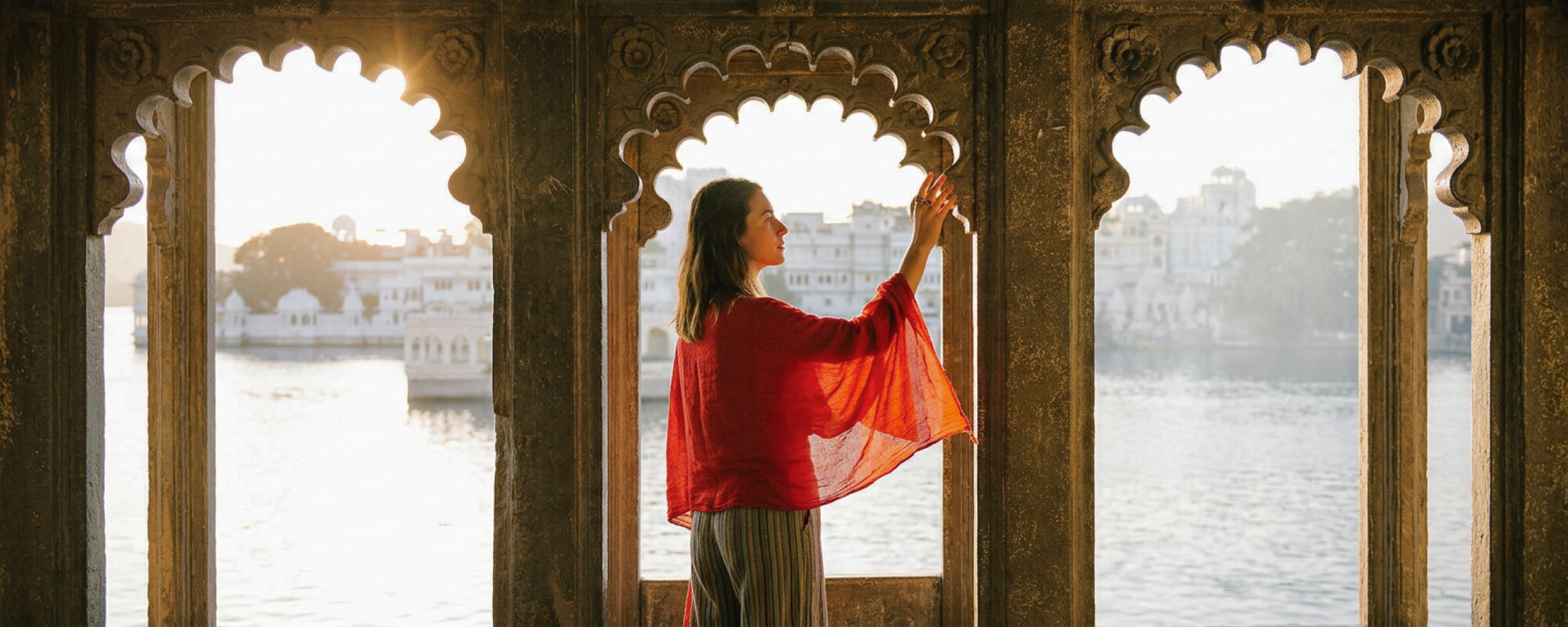 Woman in red shawl standing in ornate Indian palace window overlooking Lake Pichola in Udaipur at sunset