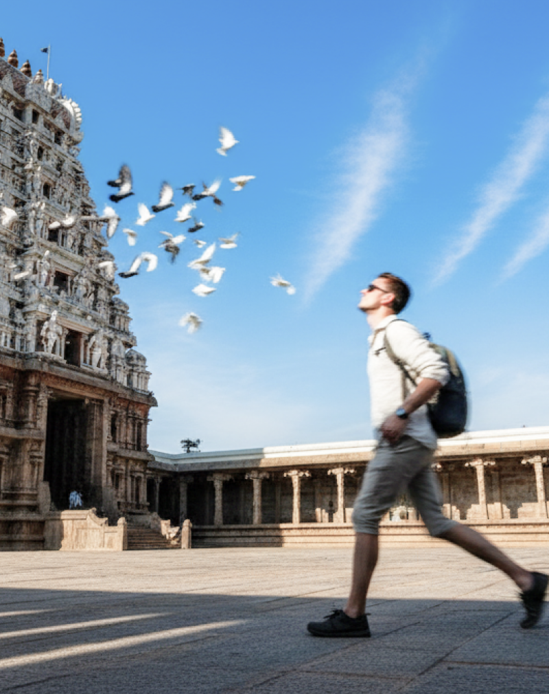 A Caucasian tourist walking through the courtyard of Kapaleeshwarar Temple in Chennai, with pigeons in flight against a vivid blue sky — Chennai travel guide thumbnail