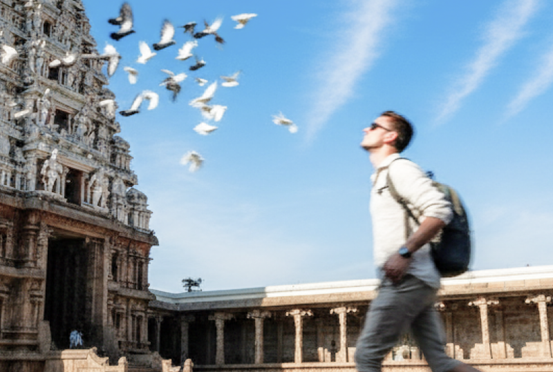 A Caucasian tourist walking through the courtyard of Kapaleeshwarar Temple in Chennai, with pigeons in flight against a vivid blue sky — Chennai travel guide thumbnail