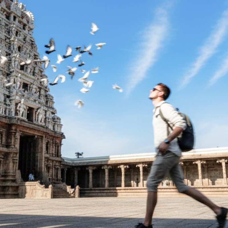 A Caucasian tourist walking through the courtyard of Kapaleeshwarar Temple in Chennai, with pigeons in flight against a vivid blue sky — Chennai travel guide thumbnail