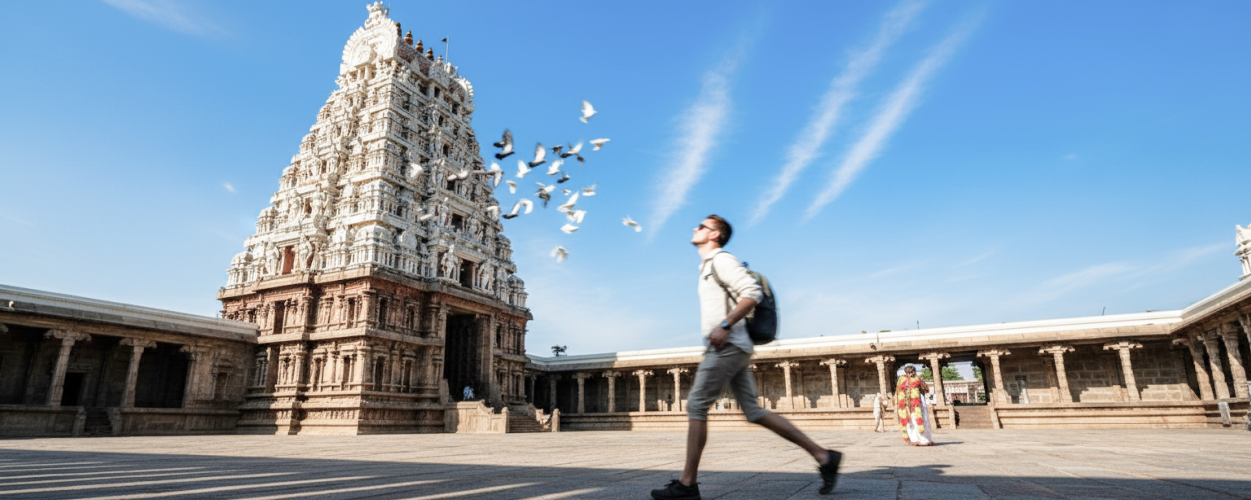 A Caucasian tourist walking through the courtyard of Kapaleeshwarar Temple in Chennai, with pigeons in flight against a vivid blue sky — Chennai travel guide