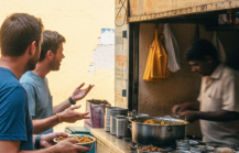 Two foreign tourists ordering misal pav at a street stall in Pune — a practical Pune food guide moment captured candidly, with a motorbike and locals passing in the background thumbnail