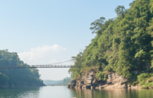A wooden boat navigating a lush green river gorge with a suspension bridge ahead, a scenic river journey to experience in India in June and July thumbnail