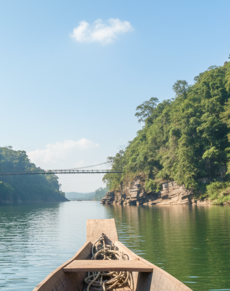 A wooden boat navigating a lush green river gorge with a suspension bridge ahead, a scenic river journey to experience in India in June and July thumbnail