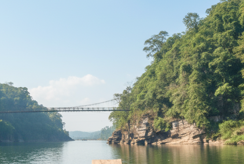 A wooden boat navigating a lush green river gorge with a suspension bridge ahead, a scenic river journey to experience in India in June and July thumbnail